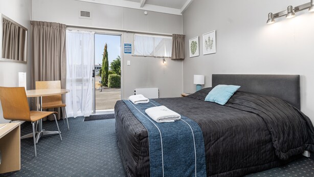 A hotel room with a bed, a desk, a chair, a window with curtains, and a door leading outside.