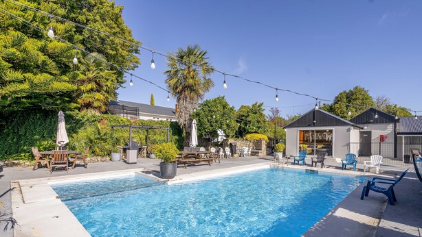 A swimming pool with lounge chairs and umbrellas, surrounded by greenery and a building with a modern design. - Amble Inn Motel - Masterton (Masterton)
