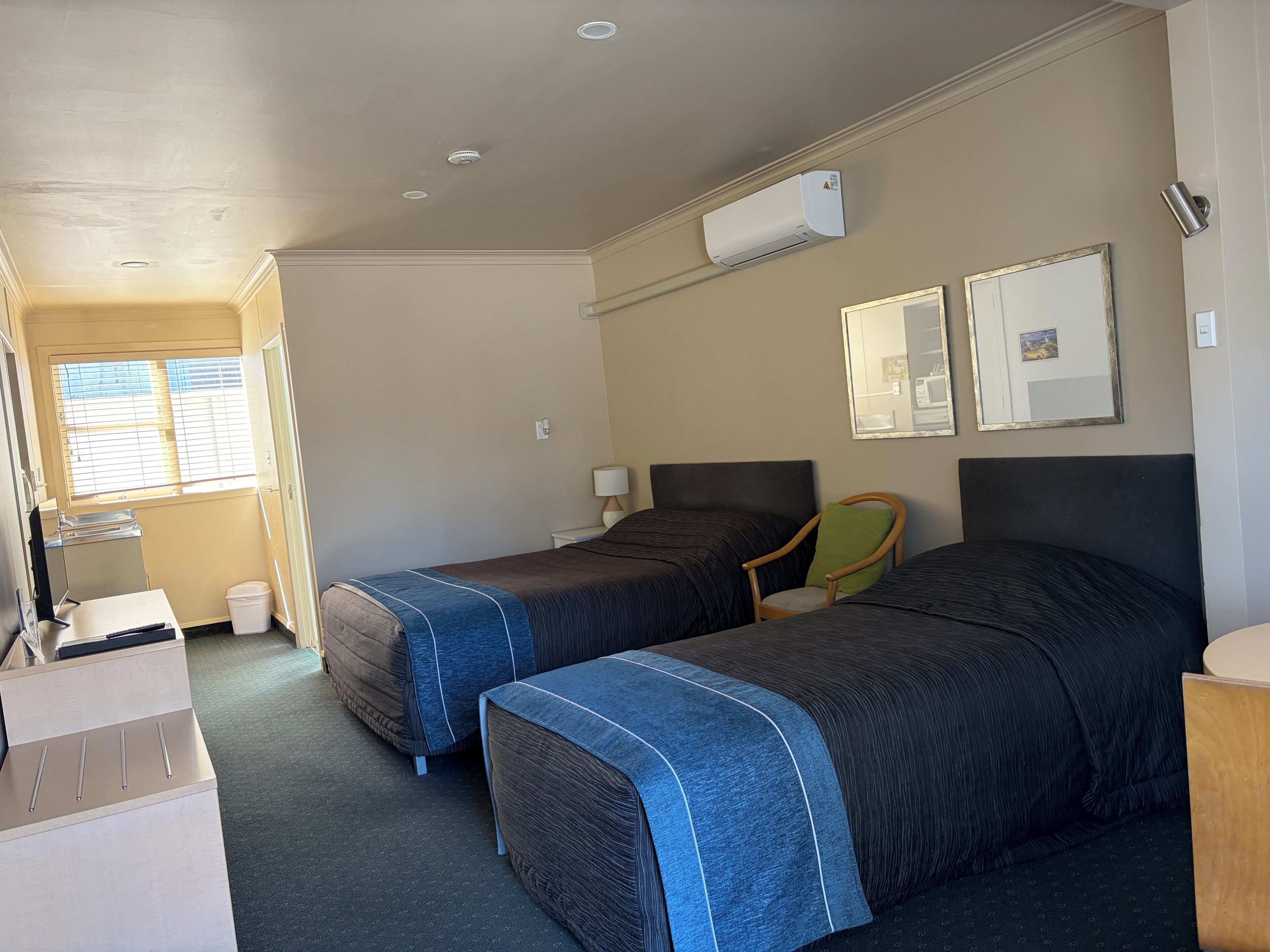 Une chambre d’hôtel avec deux lits, un bureau, une chaise, un appareil de climatisation et une fenêtre avec des stores.
