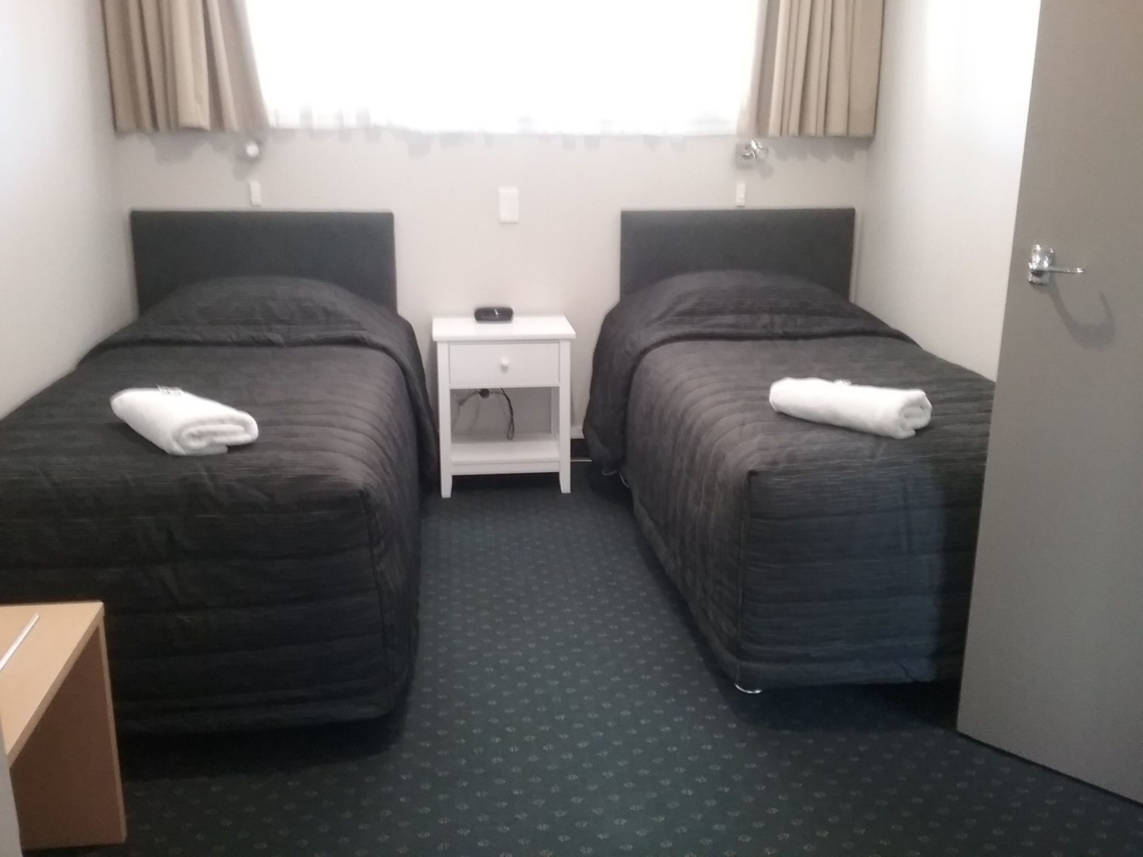 A hotel room with two single beds, a small bedside table with a lamp, a door, and a window with curtains.