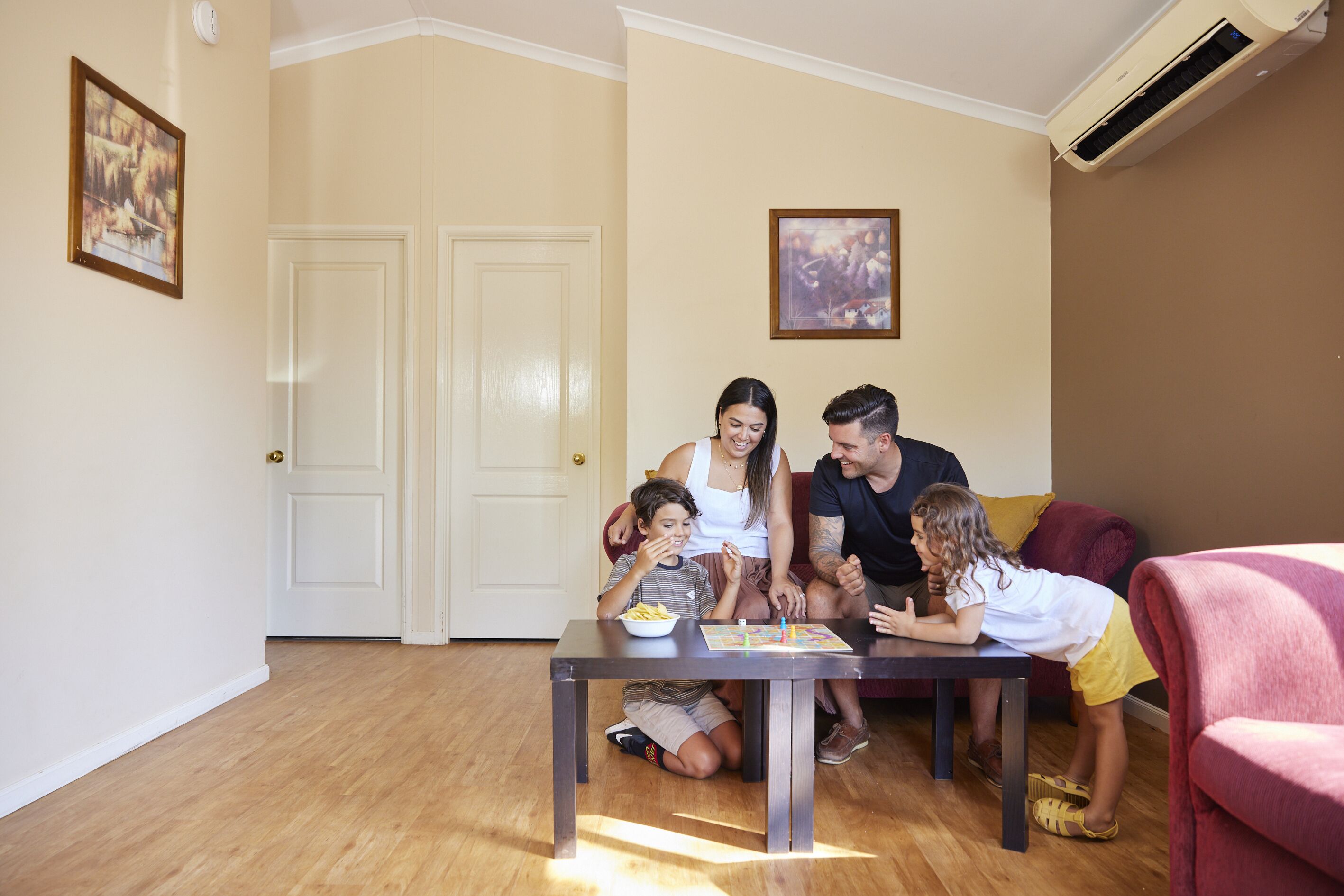 A family of four seated around a table with a bowl of snacks, in a room with wooden flooring, a door, and an air conditioning unit.