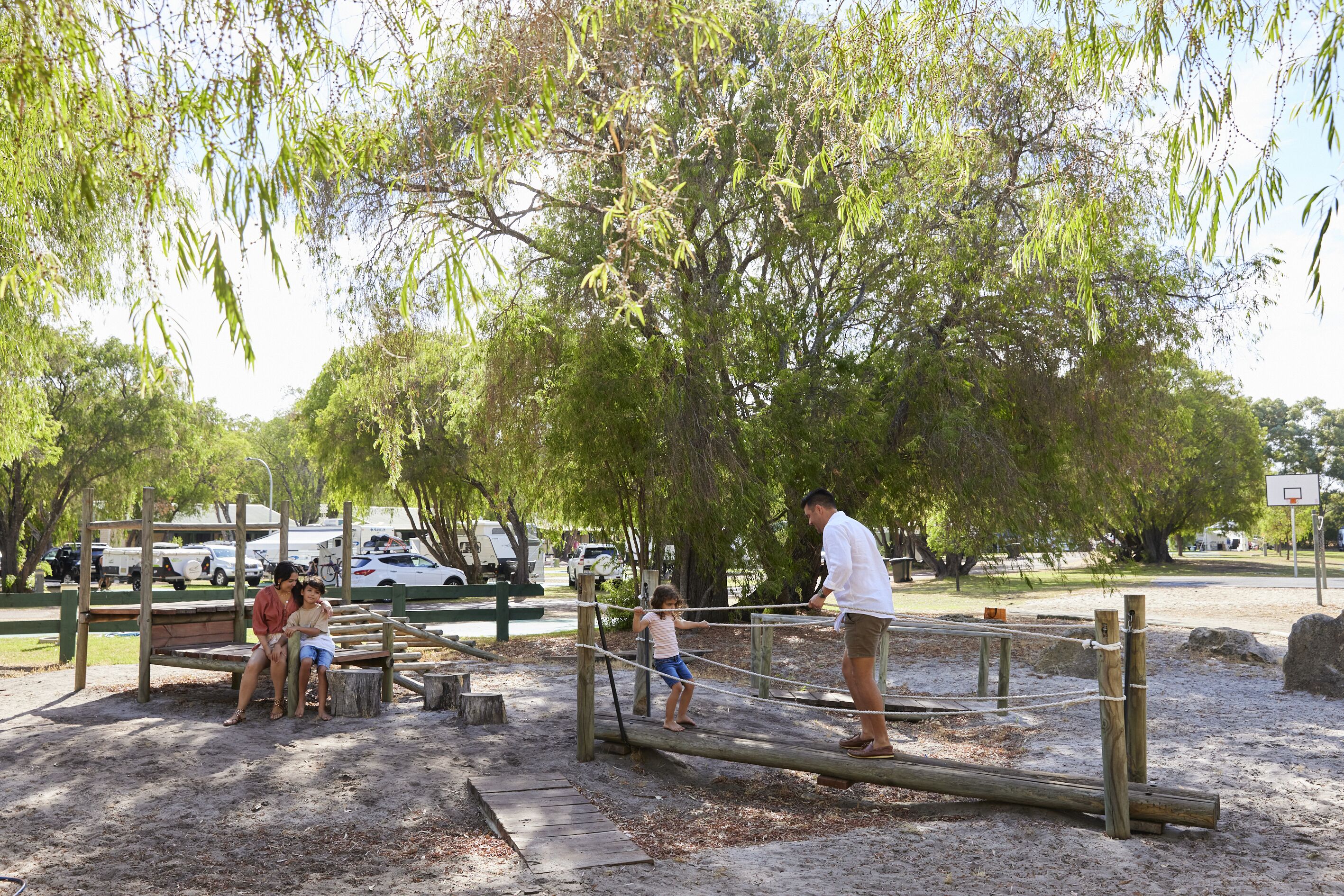 A playground with a rope bridge, sand ground, and trees in the background.