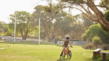 A child riding a bicycle on a grassy field with trees and benches in the background.