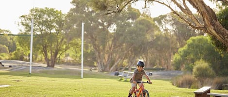 A child riding a bicycle on a grassy field with trees and benches in the background.