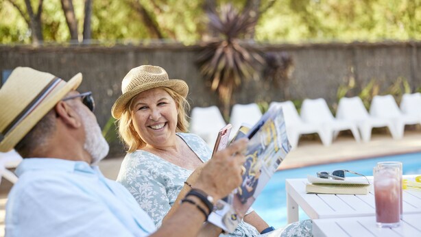 A couple sitting by a poolside lounge chair, reading a magazine. - RAC Busselton Holiday Park (Abbey)