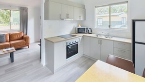 A modern kitchen with white cabinets, a built-in oven, a microwave, and a sink. There is a wooden dining table and a brown leather sofa in the adjacent living area. - RAC Busselton Holiday Park (Abbey)