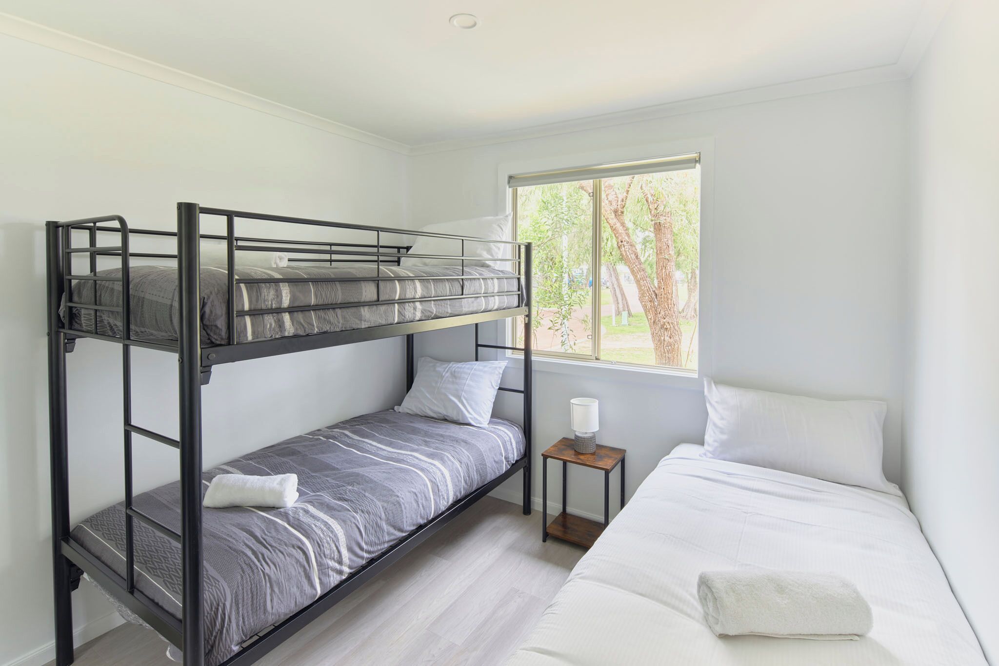 A bunk bed room with a window, a bedside table, and a white bed.