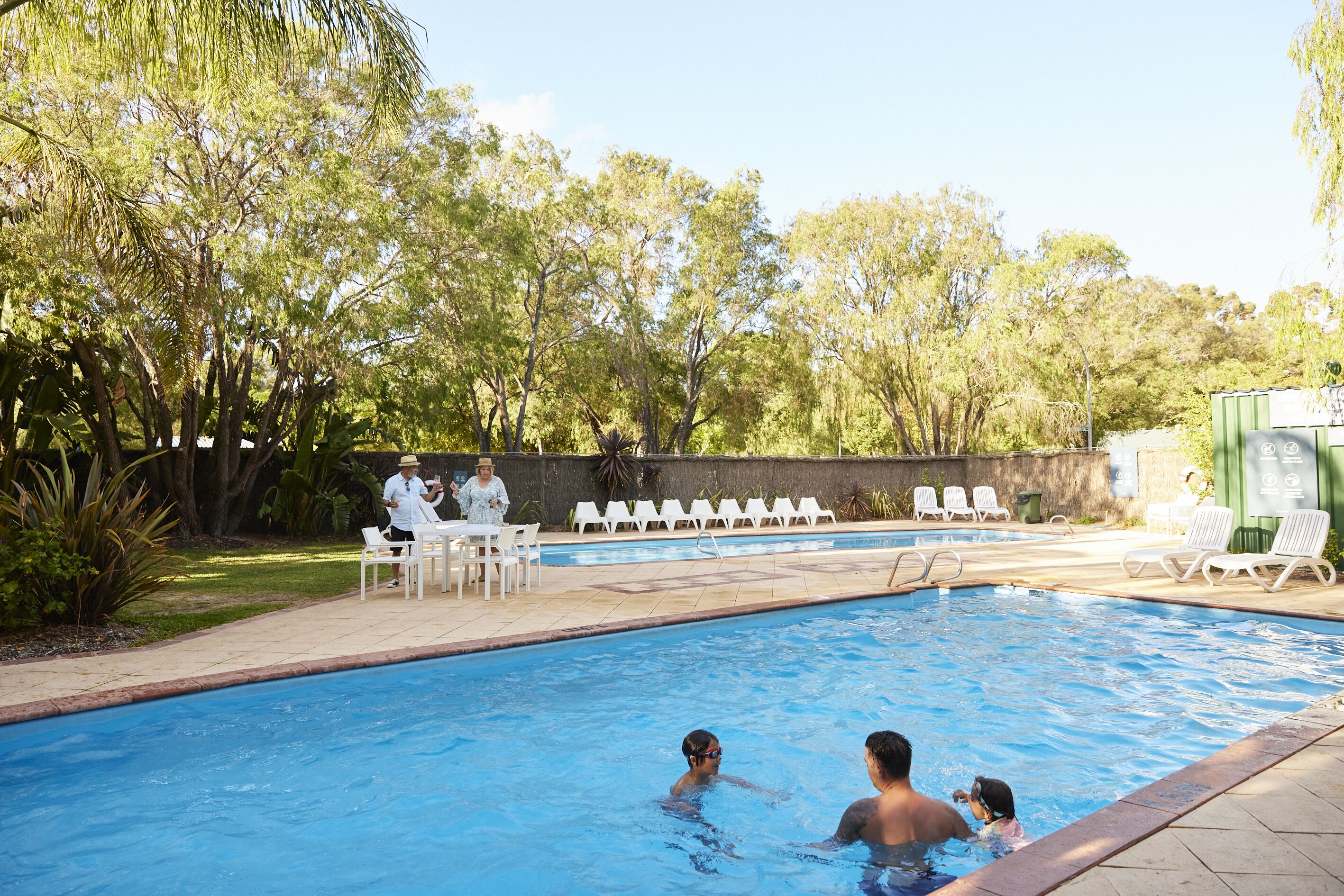 A swimming pool with people in the water, surrounded by trees and a patio area with chairs and tables.