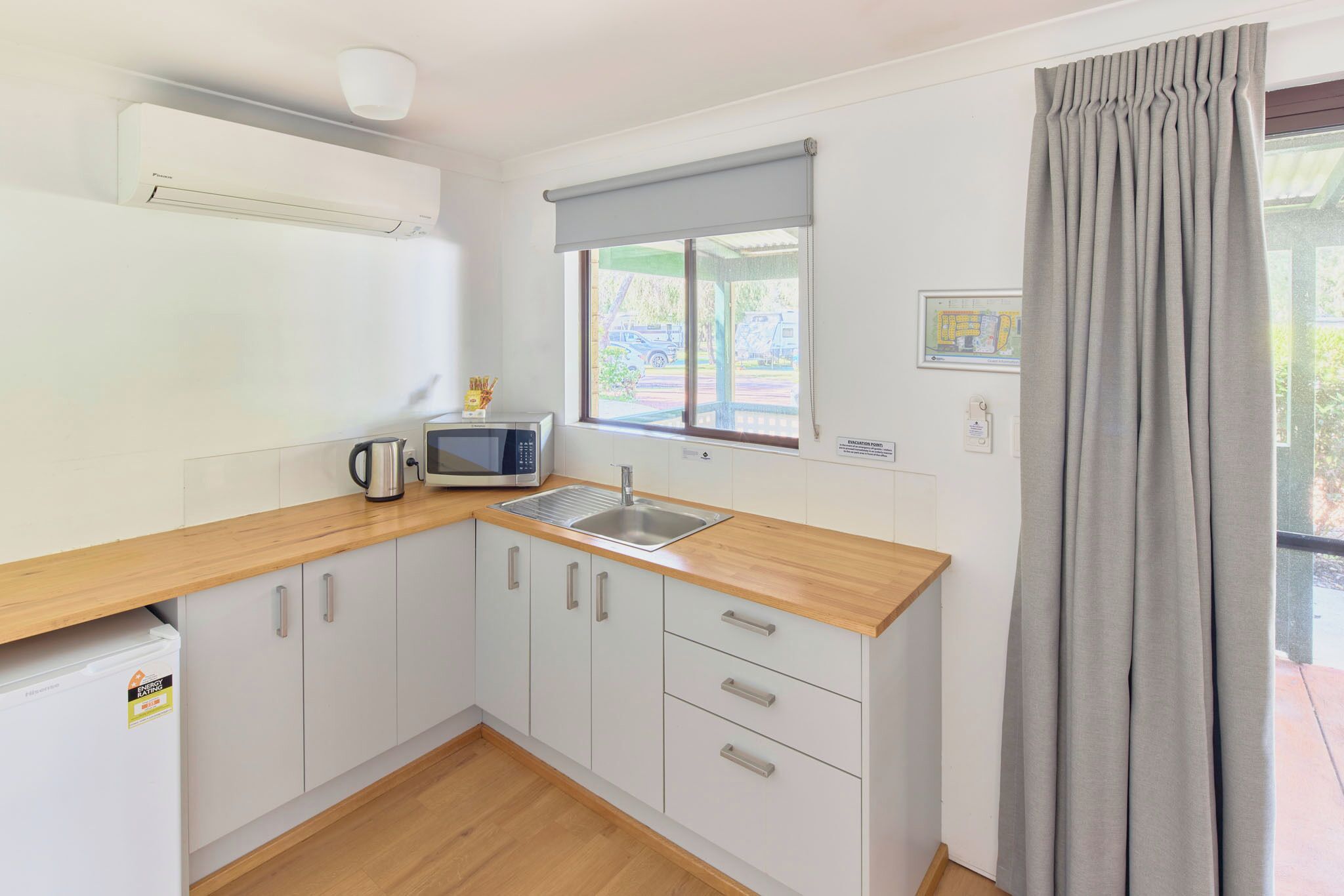 A compact kitchen with white cabinets, a wooden countertop, a microwave, a kettle, and a window with a view of the outside.