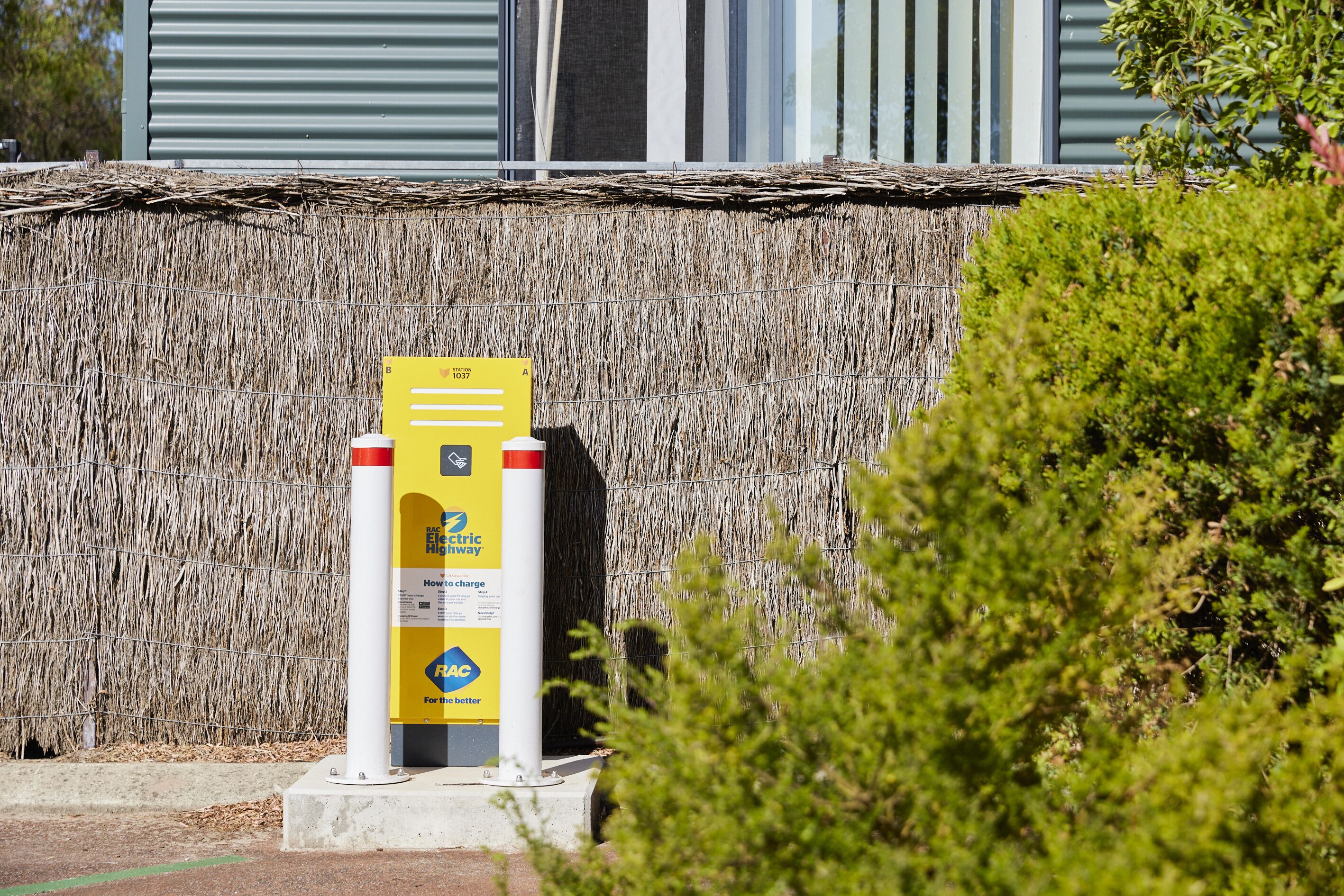 A yellow electric vehicle charging station with a sign, located in front of a building with a straw-like wall and a metal fence.
