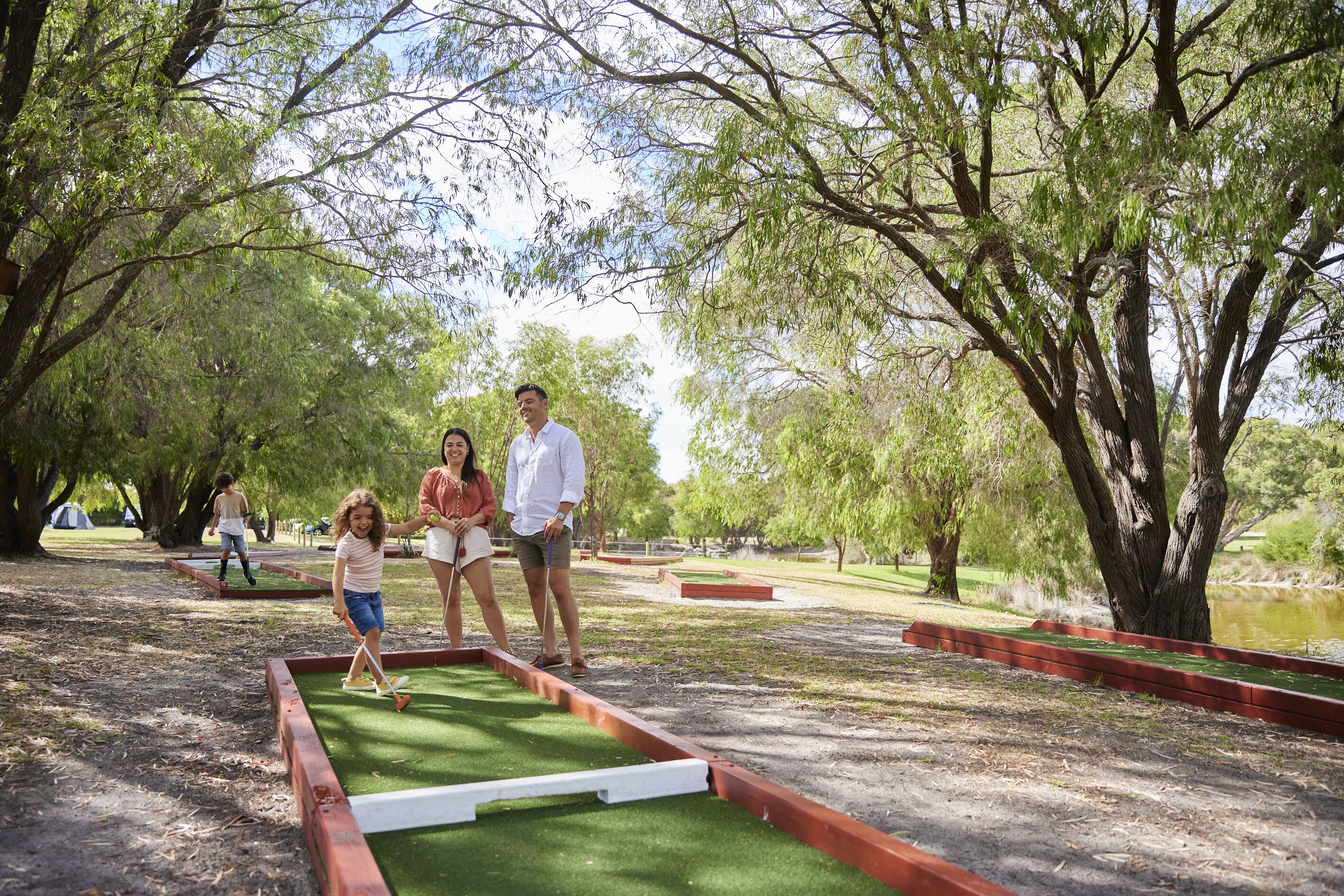 Una familia jugando minigolf en un parque con árboles y un estanque.
