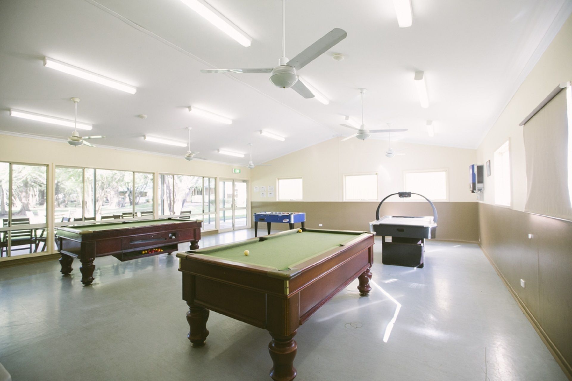 A spacious indoor pool area with multiple pool tables, ceiling fans, and large windows allowing natural light.