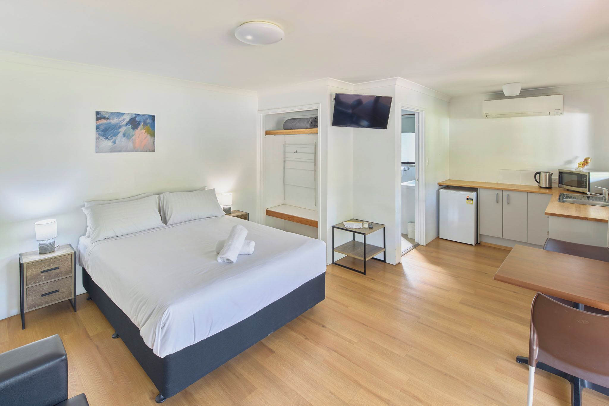 A modern hotel room with a bed, bedside table, and a small table with a lamp. There is a kitchenette with a microwave, kettle, and refrigerator.