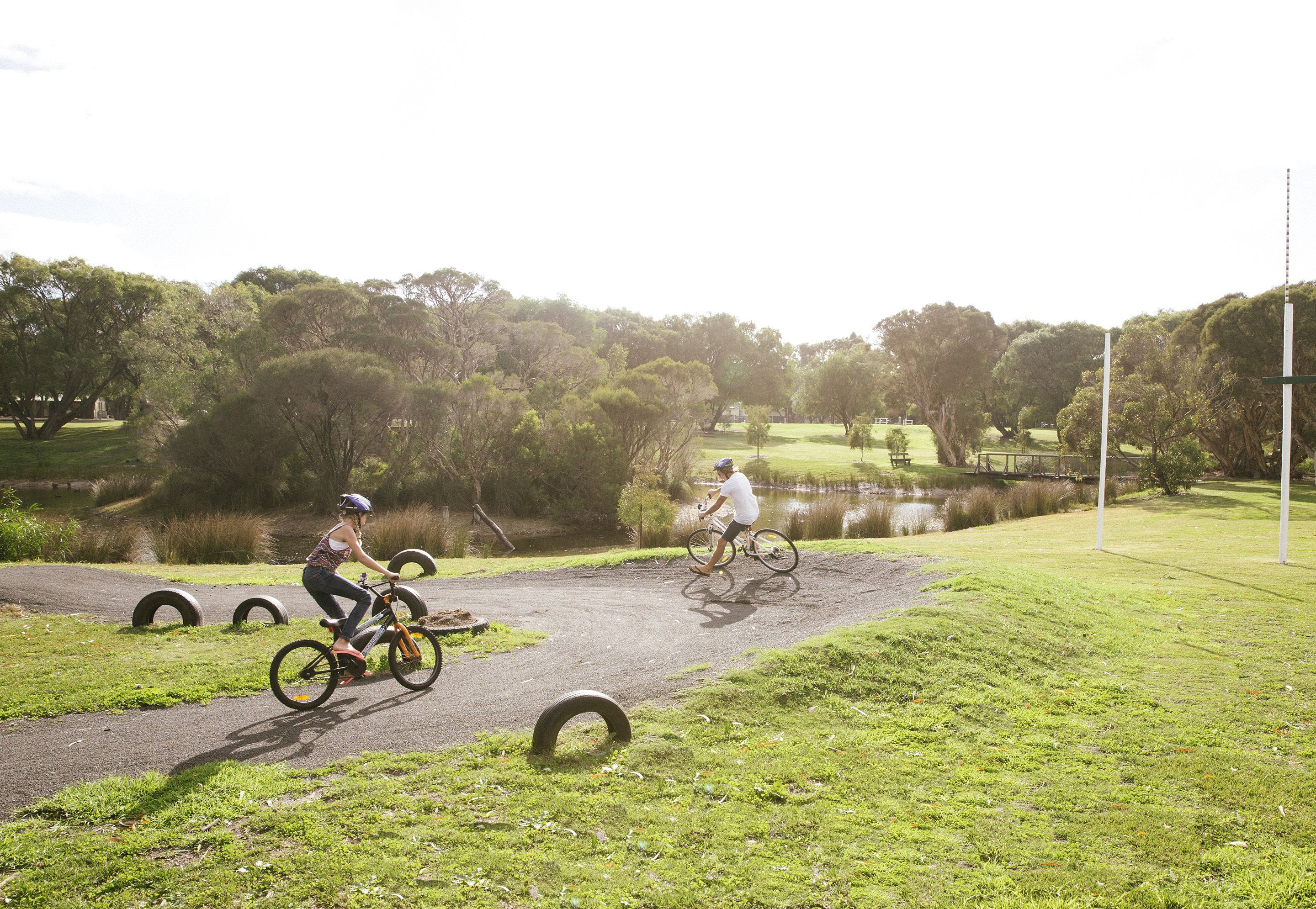 Two cyclists on a dirt path with a grassy area and trees in the background.