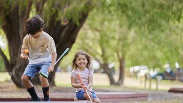 Dos niños jugando con una pelota y un bate en un parque con árboles y una estructura de ladrillo rojo.