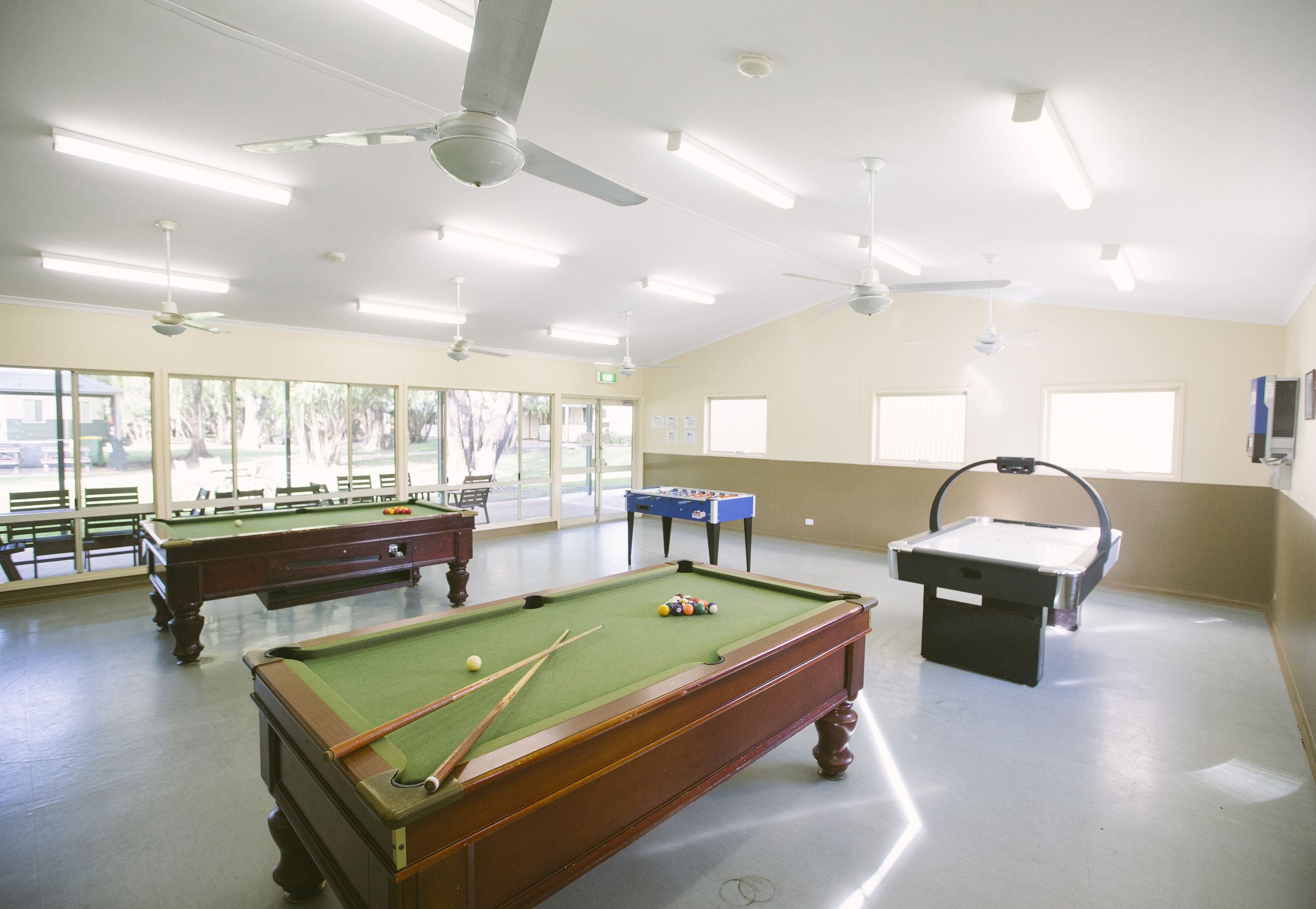 A pool room with multiple pool tables, ceiling fans, and large windows.