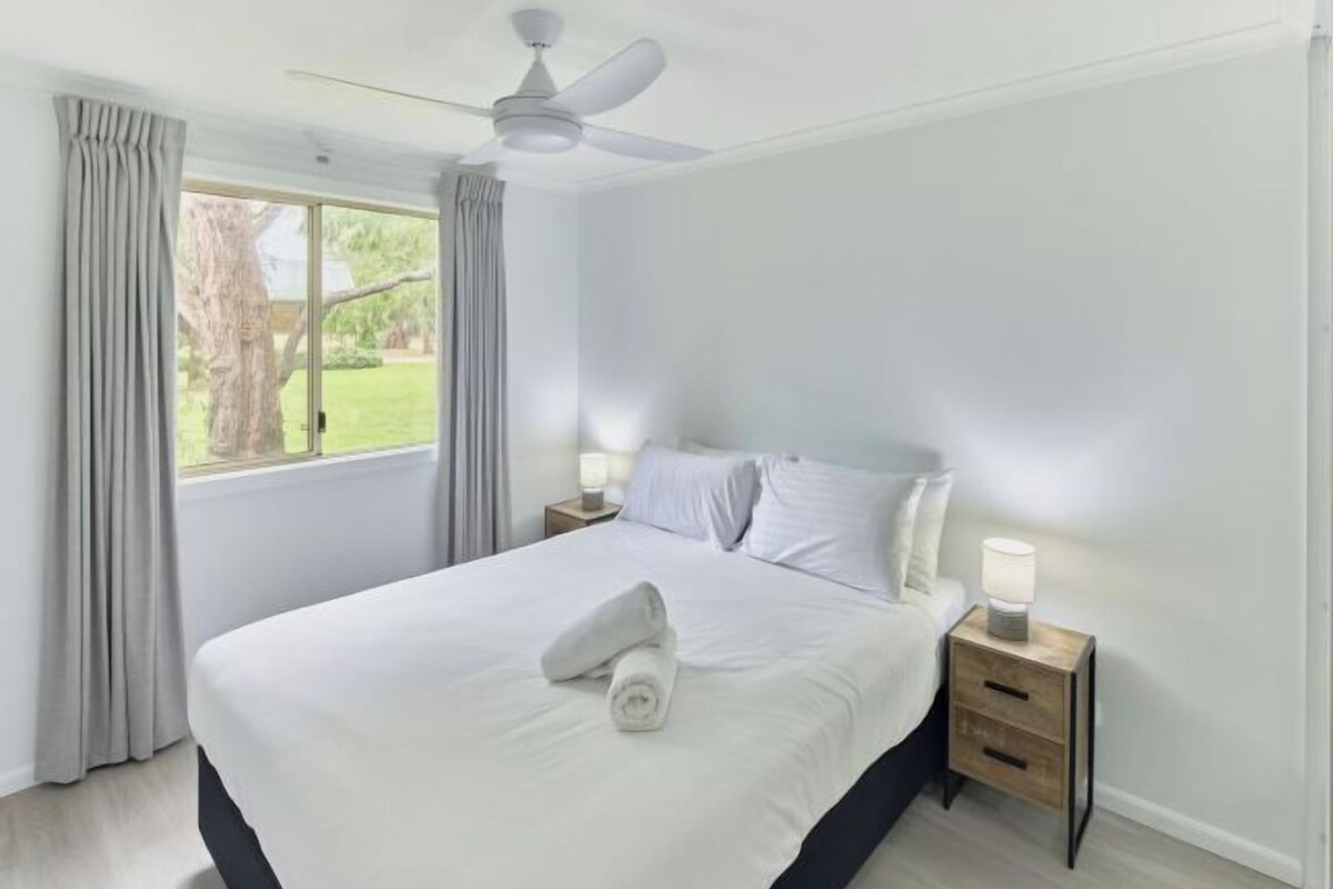 A neatly made bed with a rolled towel, a bedside table with a lamp, and a window with curtains.