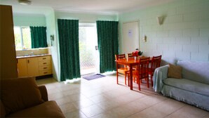 Una sala de estar con sofá, una mesa de comedor con sillas, una zona de cocina con armarios y un gran ventanal con cortinas.