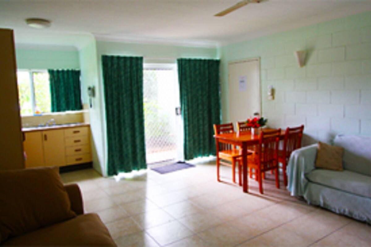A living room with a sofa, a dining table with chairs, a kitchen area with cabinets, and a sliding glass door with curtains.