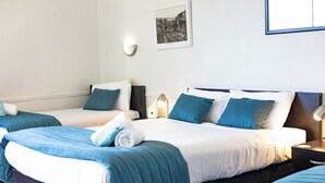 Habitación de hotel con dos camas, cada una con ropa de cama azul y almohadas blancas. Hay un cuadro enmarcado en la pared y una lámpara fijada a la pared.