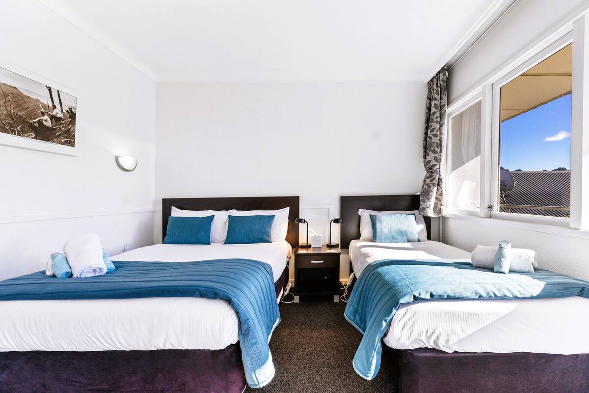 A hotel room with two beds, a nightstand, a wall-mounted light, and a window with a view.