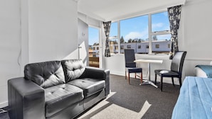 Una habitación de hotel moderna con un sofá de cuero negro, un escritorio pequeño con una silla, una cama y ventanas grandes con vistas a otros edificios.