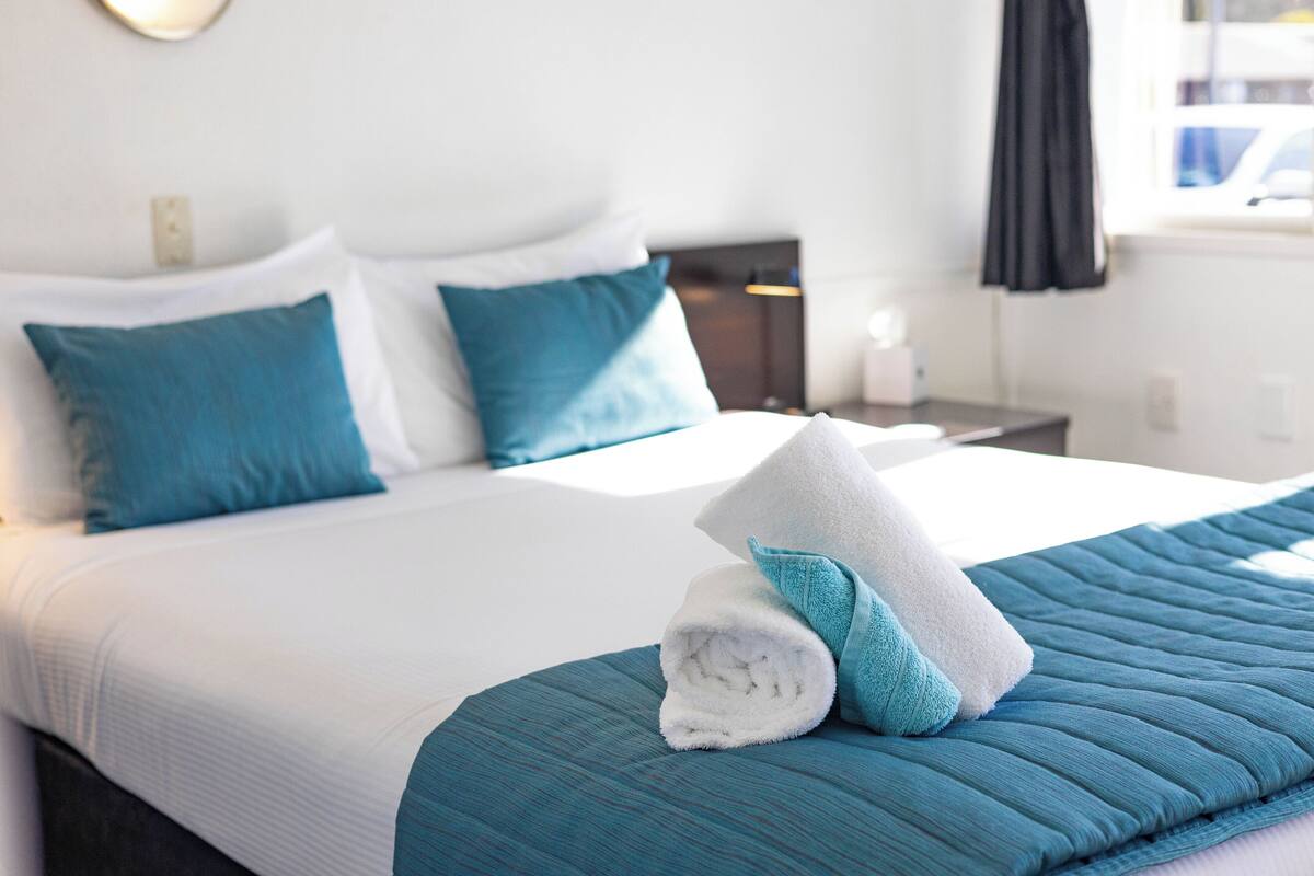 A neatly made bed with blue and white bedding, a rolled towel, and a teal throw blanket.
