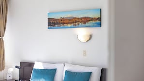 Una habitación de hotel con cama, mesita de noche, lámpara, un cuadro de montaña y lago, y un aplique de pared.
