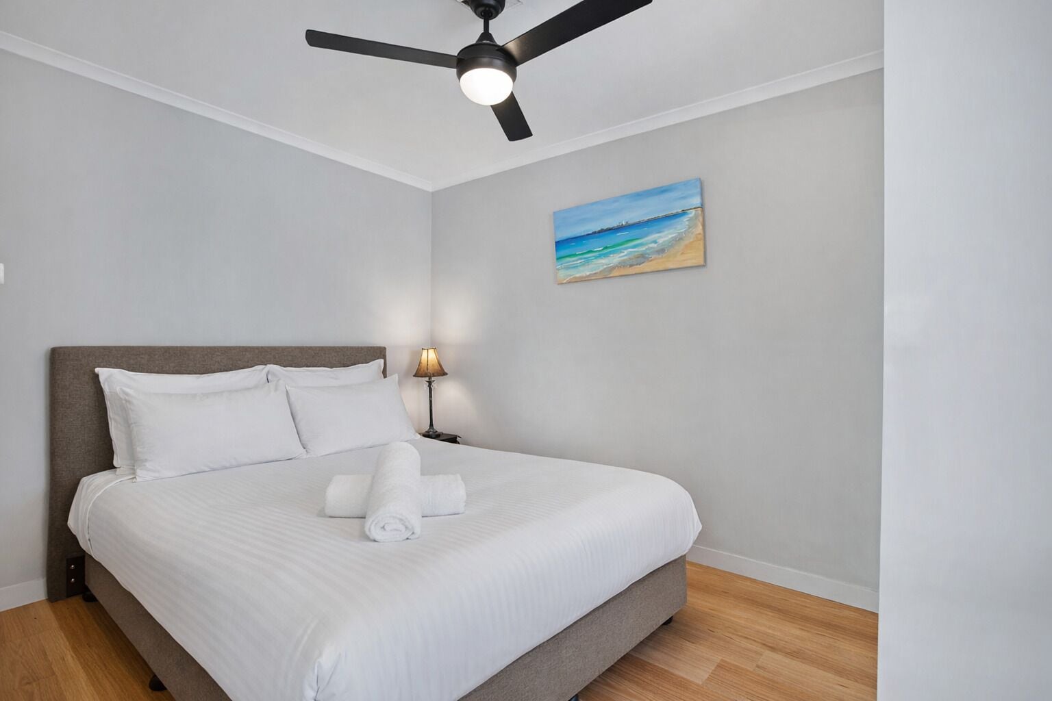 Een net opgemaakt bed met witte lakens en een opgerolde handdoek, een plafondventilator, een nachtkastje met een lamp, en een schilderij van een strandlandschap aan de muur.