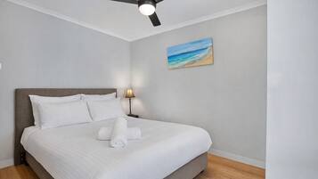 Een net opgemaakt bed met witte lakens en een opgerolde handdoek, een plafondventilator, een nachtkastje met een lamp, en een schilderij van een strandlandschap aan de muur.
