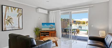 A modern living room with a black leather sofa, a wooden coffee table, and a TV on a wooden stand. There is a sliding glass door leading to an outdoor area with a pool and chairs.