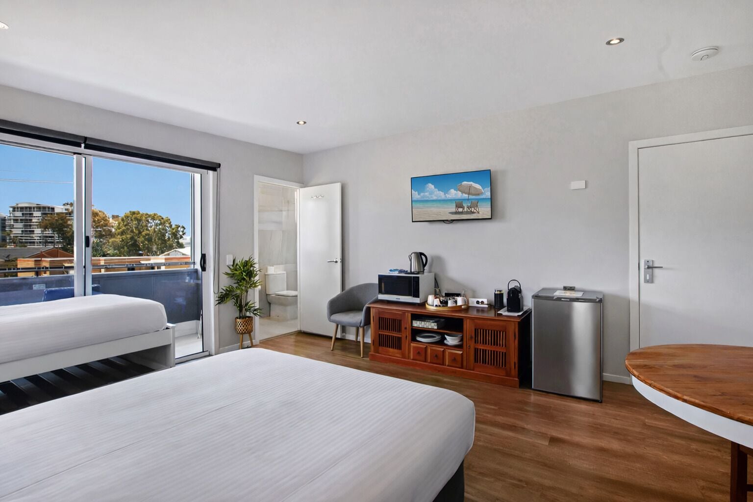 Una habitación de hotel moderna con dos camas, una zona de comedor, un televisor, un refrigerador pequeño y vistas a la ciudad.