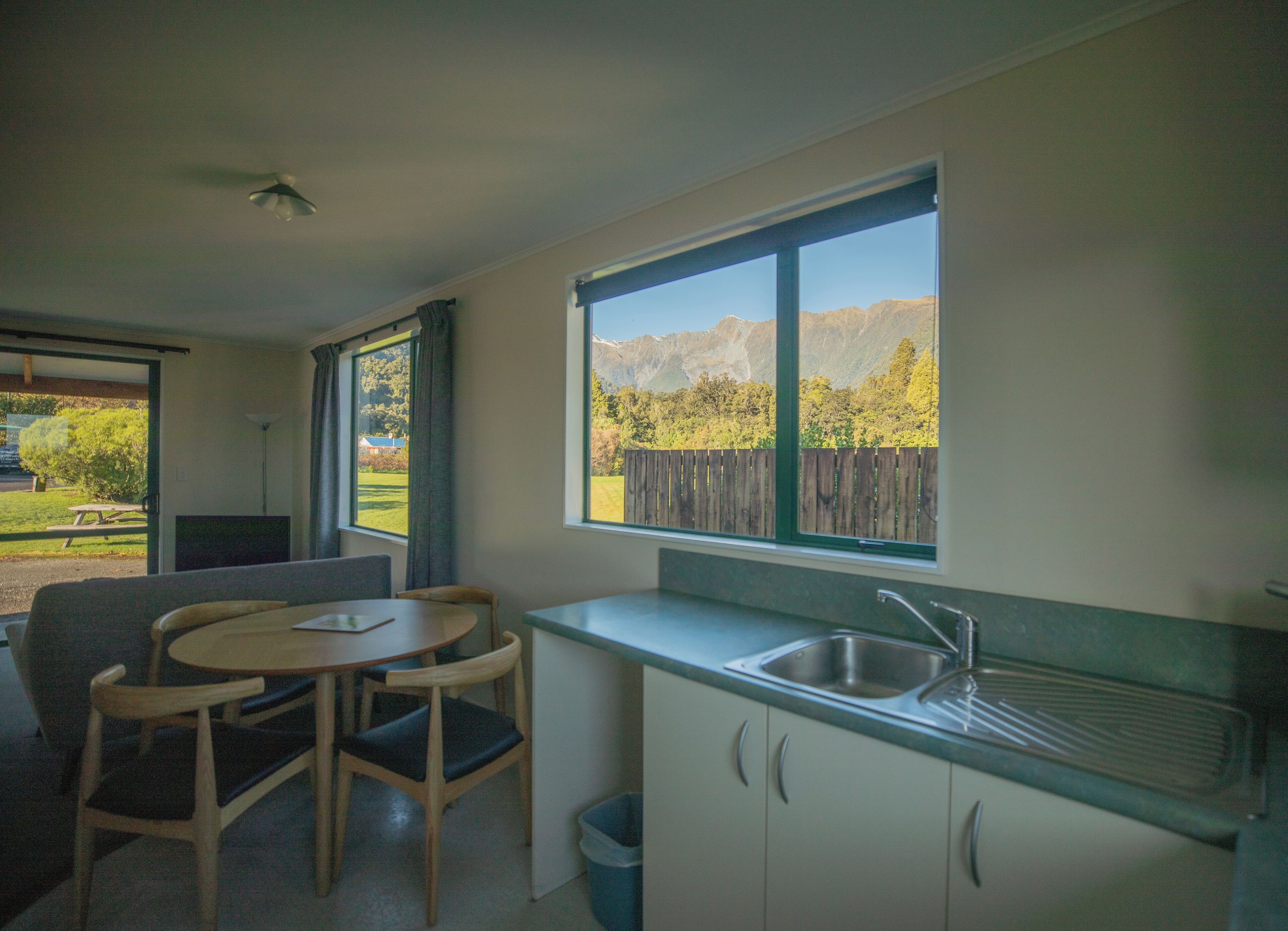 A compact kitchen with a sink, a dining table with chairs, and a view of a mountainous landscape through a large window.