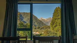 A room with a large window offering a view of a mountainous landscape, a wooden table, and a chair. - Rainforest Motel (Fox Glacier)
