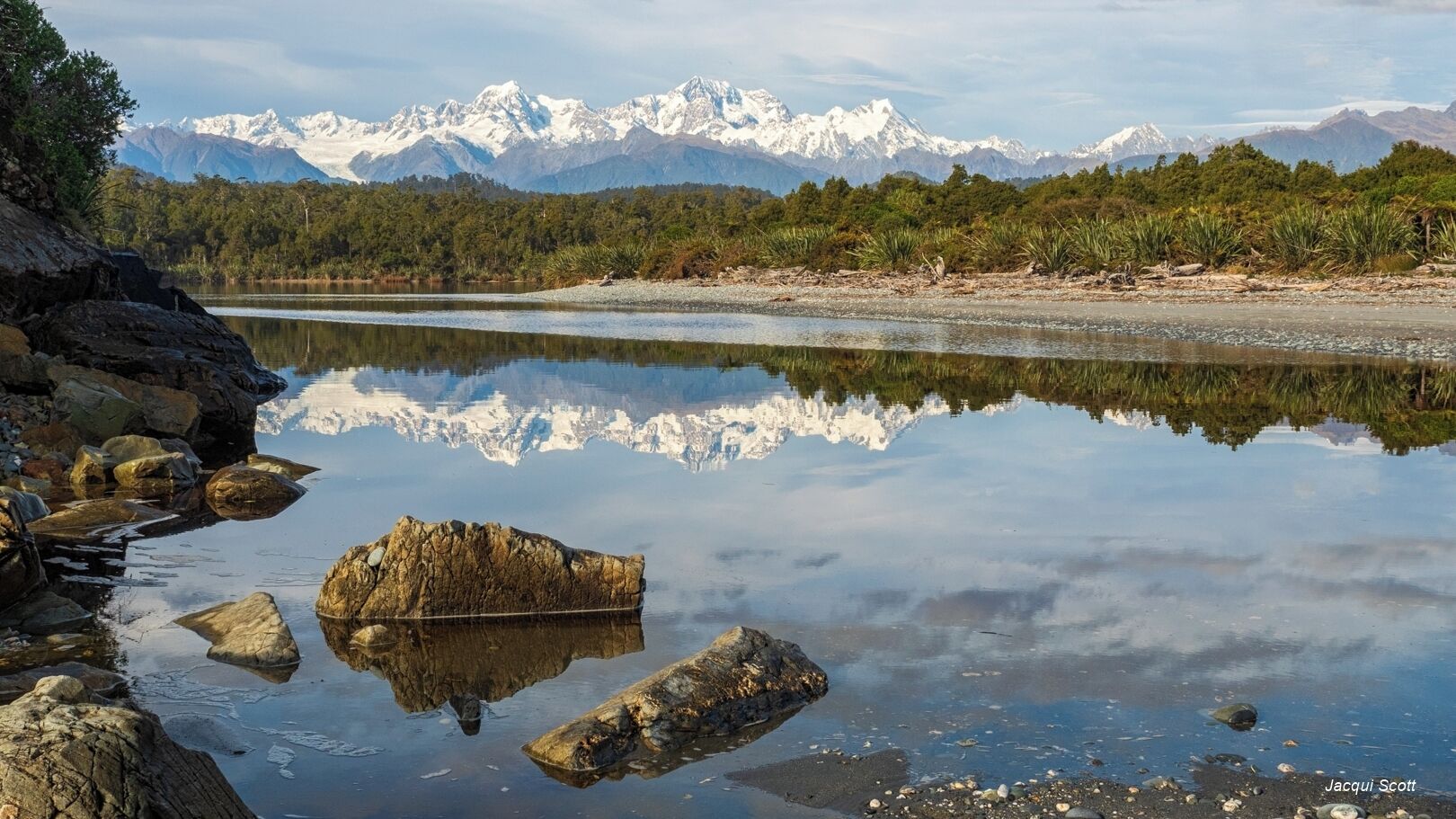 A calm lake with a rocky shoreline and a reflection of snow-capped mountains in the background.