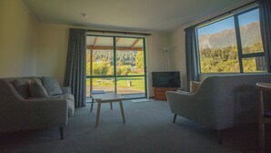 A living room with two sofas, a TV, and a large window offering a mountain view. - Rainforest Motel (Fox Glacier)