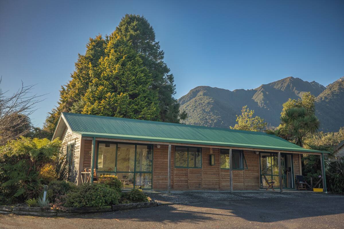 A wooden cabin with a green roof, large windows, and a gravel driveway.