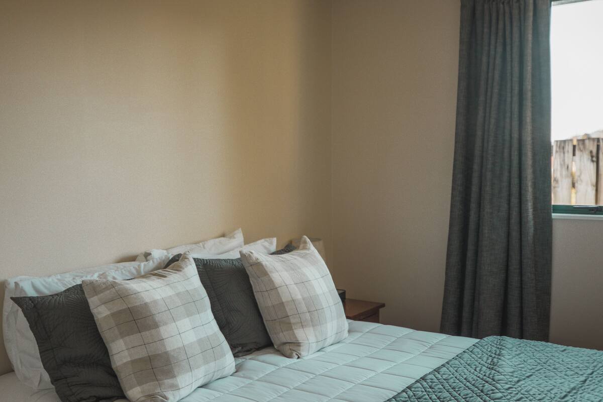 A neatly made bed with a quilted cover and several pillows, a bedside table, and a window with curtains.