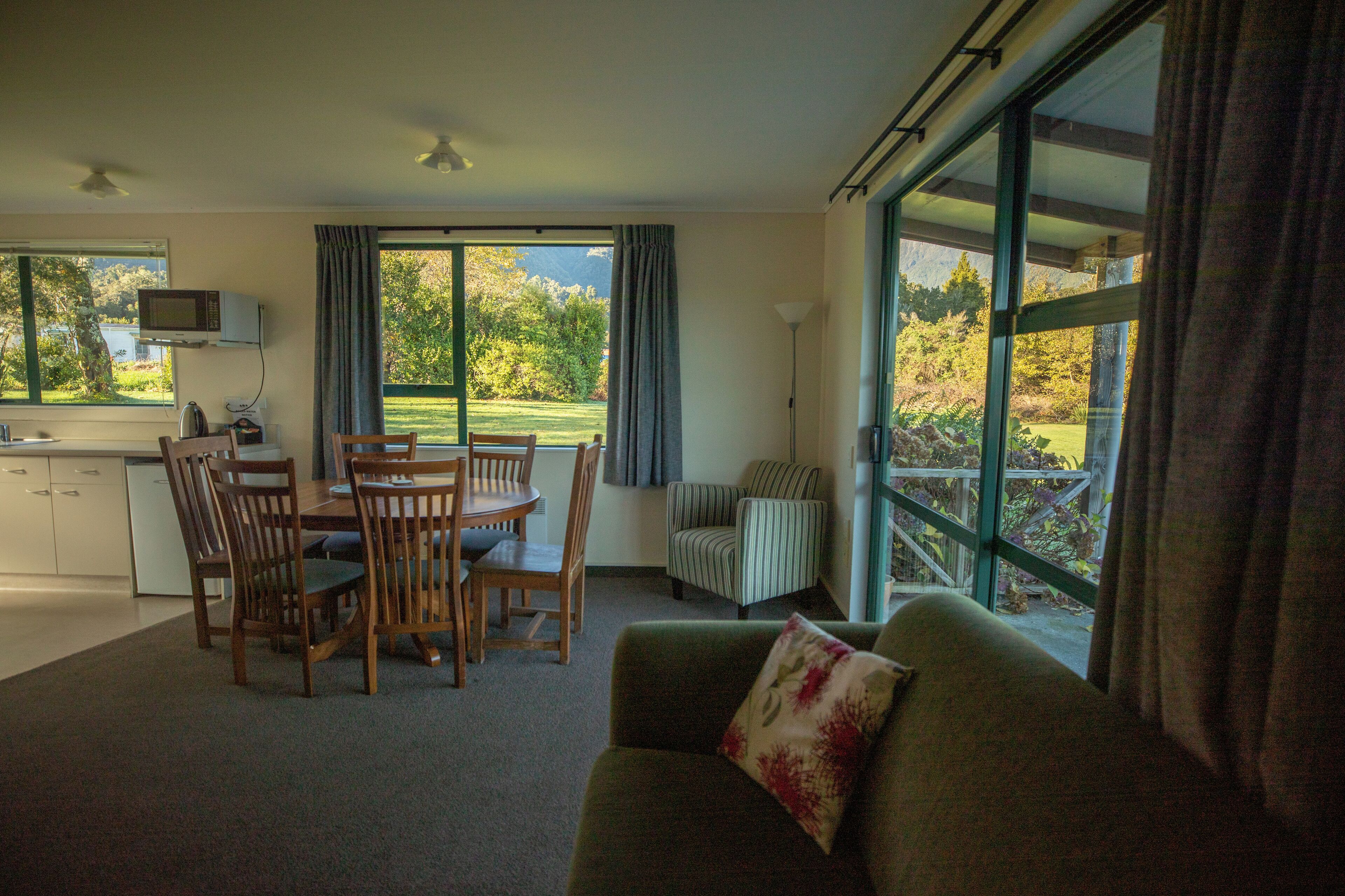 A room with a dining area, a sofa, and a large window with a view of a garden and mountains.