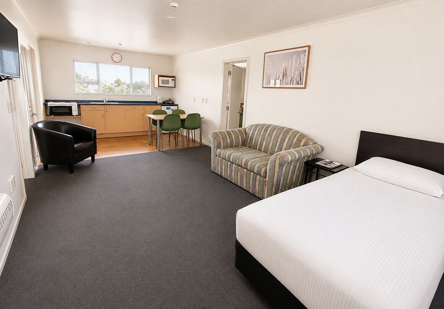 A hotel room with a bed, a sofa, a dining table with chairs, a kitchen area, and a view of the outside.