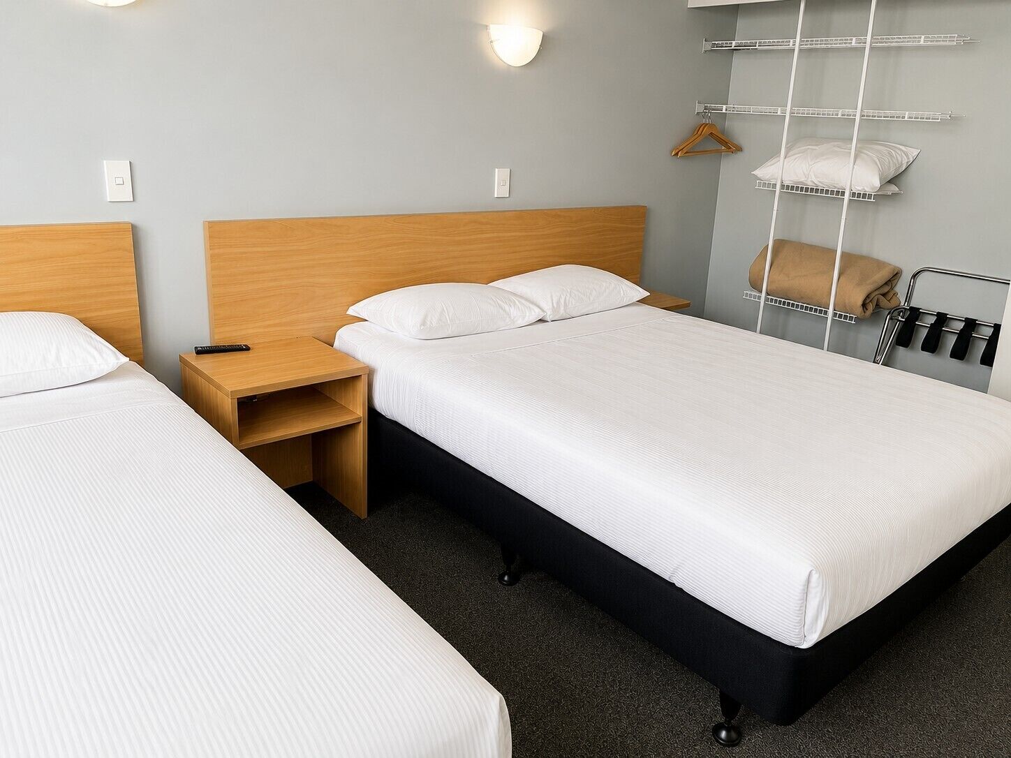 A hotel room with two single beds, a wooden nightstand, a wall-mounted shelf with a towel, and a hanging coat rack.