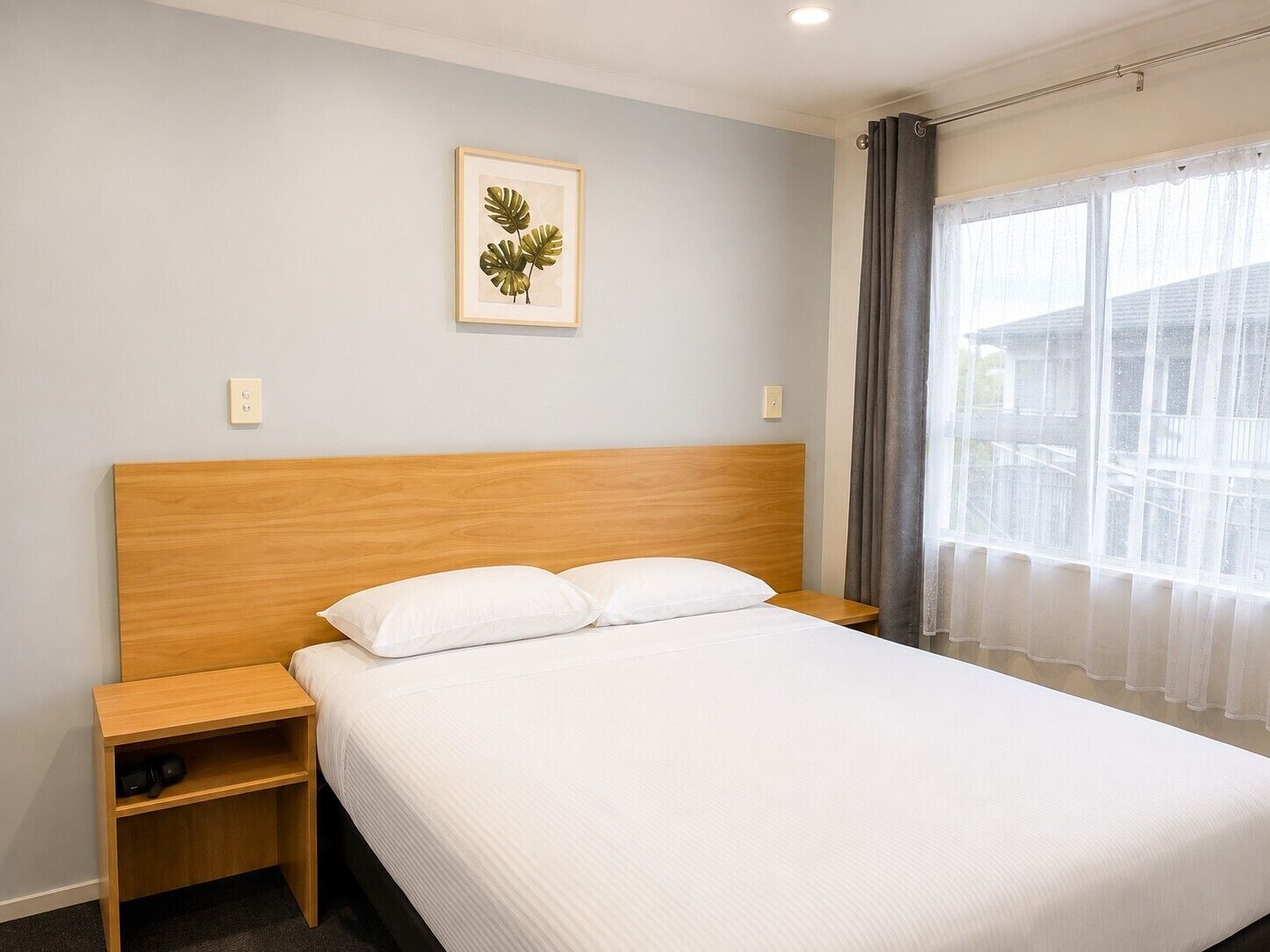 A hotel room with a wooden headboard, a single bed with white bedding, a nightstand, and a framed plant on the wall.