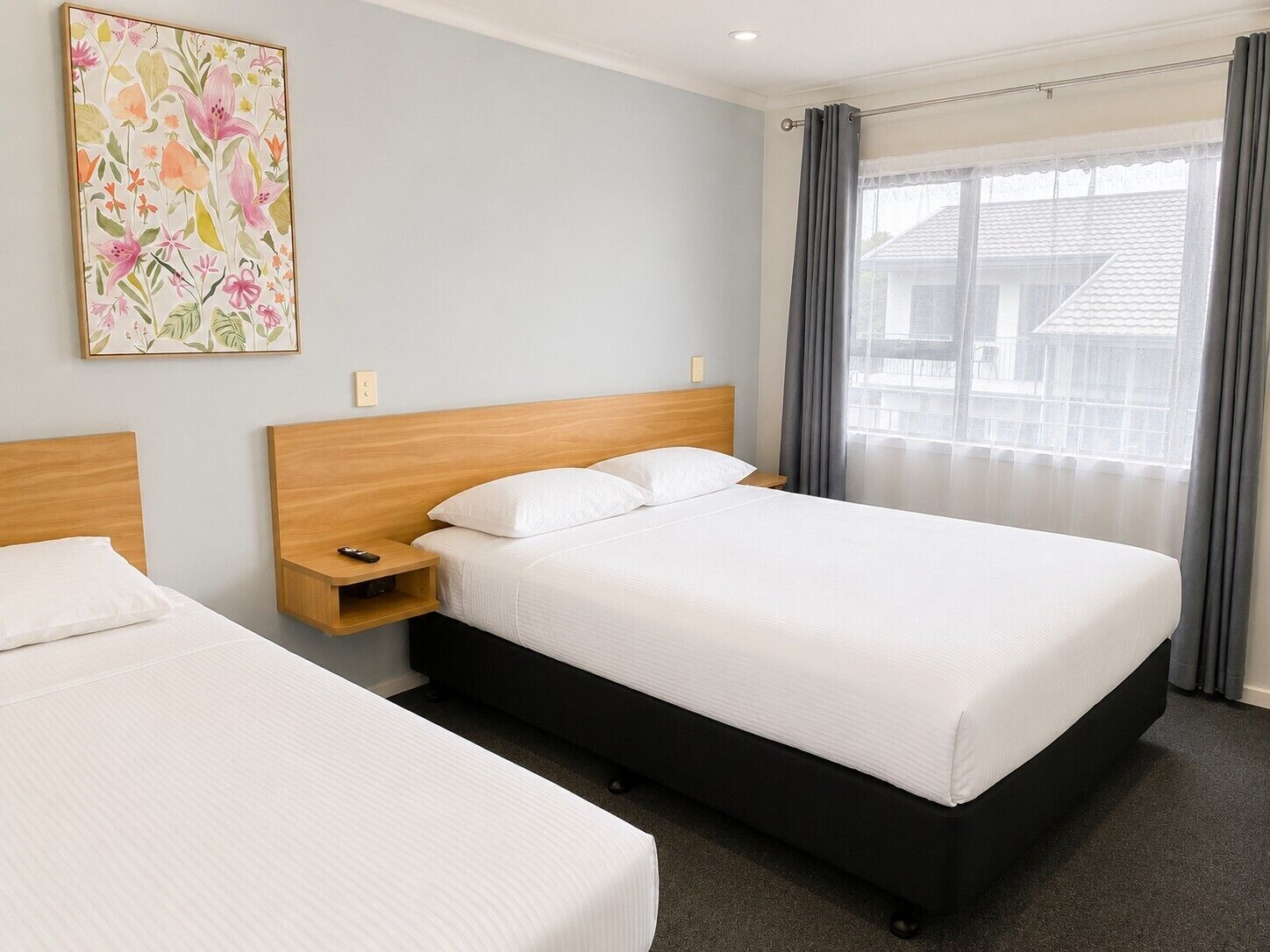 A hotel room with two beds, a wooden headboard, a bedside table with a remote control, and a window with curtains.