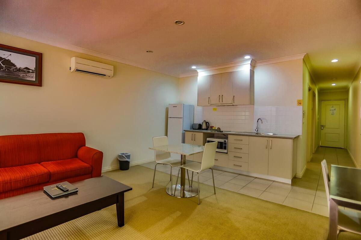 A compact living space with a red sofa, a dining table with chairs, a kitchen area with cabinets and a refrigerator, and a hallway leading to another room.