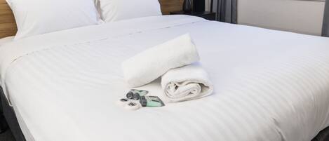 Una cama bien hecha con sábanas blancas, una toalla enrollada y artículos de aseo sobre una superficie limpia.