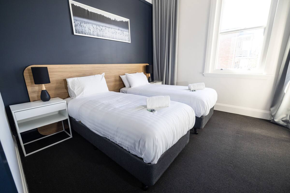 A hotel room with two beds, a nightstand with a lamp, a framed picture on the wall, and a window with curtains.