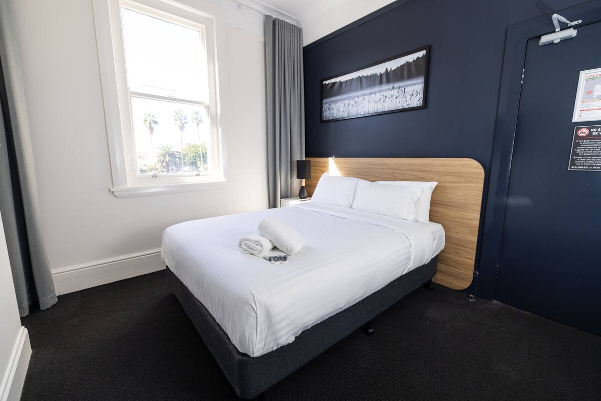 A neatly made bed with white linens and a rolled towel, a nightstand with a lamp, a framed picture on the wall, and a window with curtains.