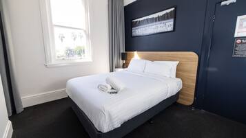 A neatly made bed with white linens and a rolled towel, a nightstand with a lamp, a framed picture on the wall, and a window with curtains.