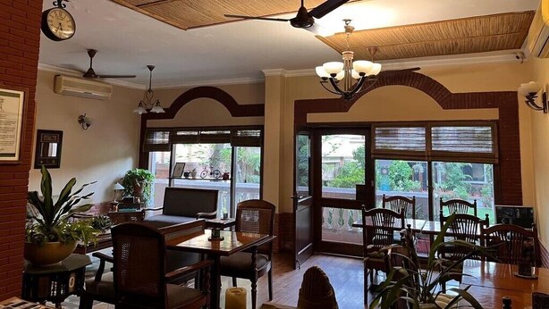 A dining area with a table and chairs, a ceiling fan, and large windows with a view of greenery outside. - The Estate Villa (New Delhi)