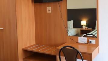 A hotel room with a wooden desk, a chair, a flat-screen TV mounted on the wall, and a bedside table with a lamp.